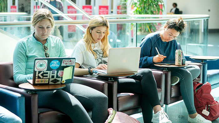 Three students seated in the Library in lounge chairs, studying on their laptop computers and devices.