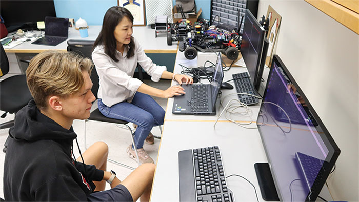 Two people in a workspace with computers and electronics; a woman is working on a laptop while a man observes. Robotics equipment and other devices are visible in the background.