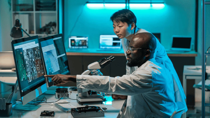 Two SDSU researchers in lab coats analyzing circuit board images on computer screens in a high-tech laboratory, with a microscope and electronic components on the desk.