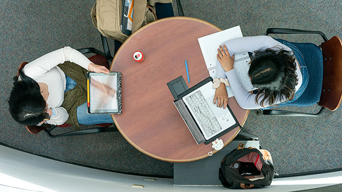 An overhead view of two students studying at a table, one at a laptop, the other on a tablet
