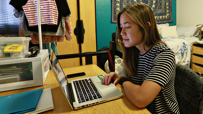 A student in her dorm room at her desk takes an online survey on her laptop