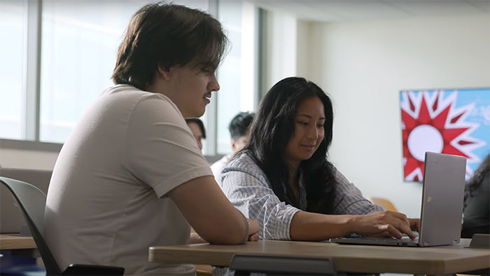 Two students sitting at a classroom table working together on a laptop, with a bright window and poster in the background.